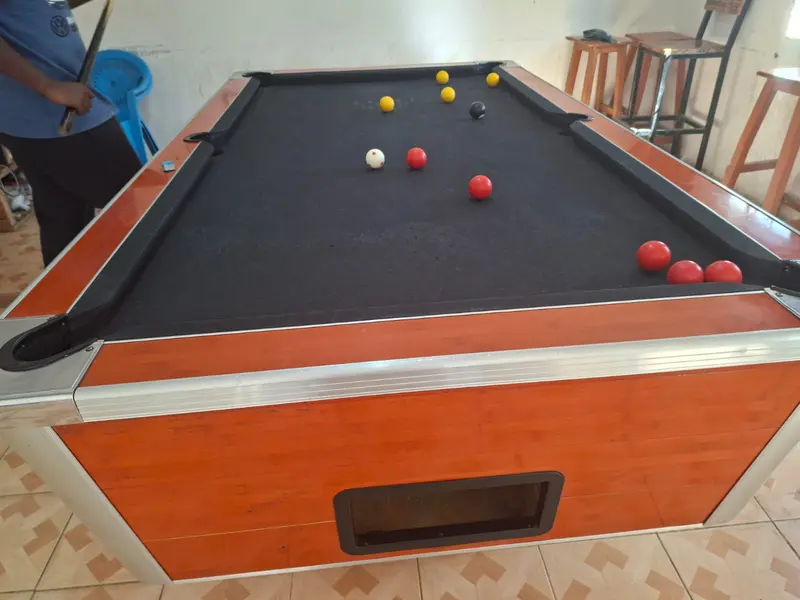 A black-cloth pool table mid-frame with red and yellow balls and the 8-ball, and a player waiting with a cue.