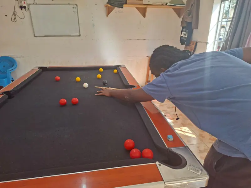 A player in a blue shirt bridges on a black-cloth pool table with reds, yellows and the 8-ball spread across the table.