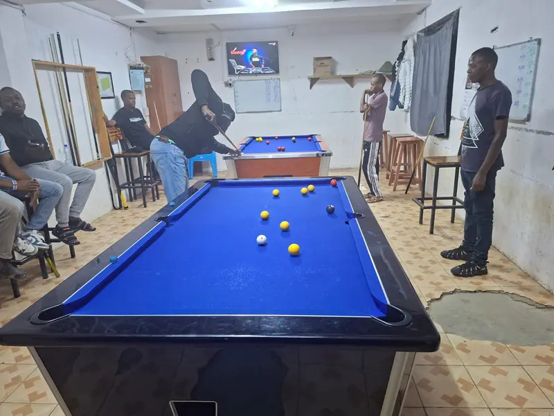 A player bends over a blue-cloth pool table at Sikiliza Piranhas Arena to take a shot while other players wait and watch, with a second table in play behind them.